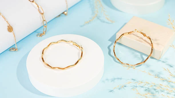 Gold-plated decorative hoop earrings displayed on minimalist white stands against a light blue background.